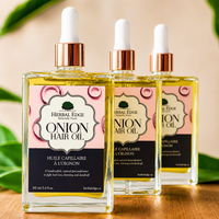 Three bottles of Herbal Edge Naturally Hair Onion Hair Oil arranged on a wooden surface with soft-focus green leaves in the background. The bottles are clear glass with rose gold droppers and pink and black labels.