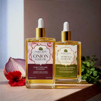 Two Herbal Edge hair oil bottles on a wooden surface, one labeled Onion Hair Oil with a purple label and one labeled Moringa Hair Oil with a green label, styled with an onion, red flower, and herbs.